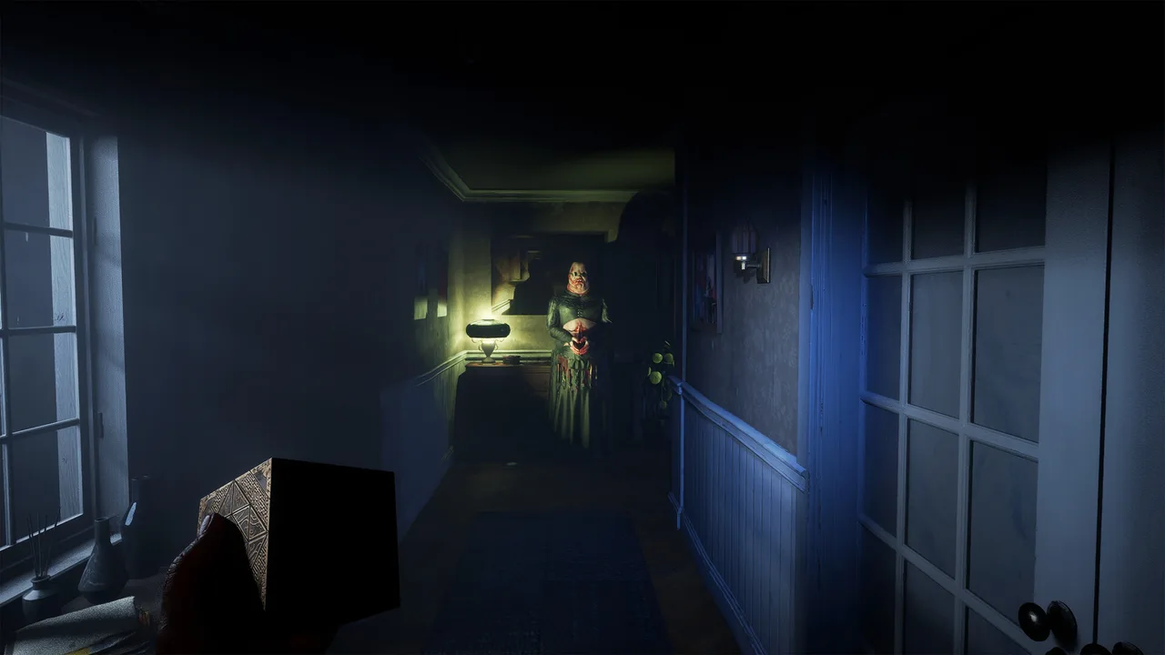 A dim hallway with patterned wallpaper, a lamp on the wall and a figure waiting at the far end